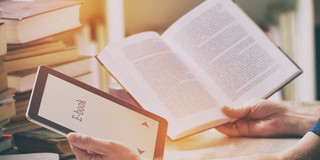 Man sitting at table with e-book reader and book A man sits with an e-book reader and a book in his hands at a table with books, stationery and papers