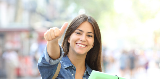 A happy student with booklets in her arm gives the thumbs up